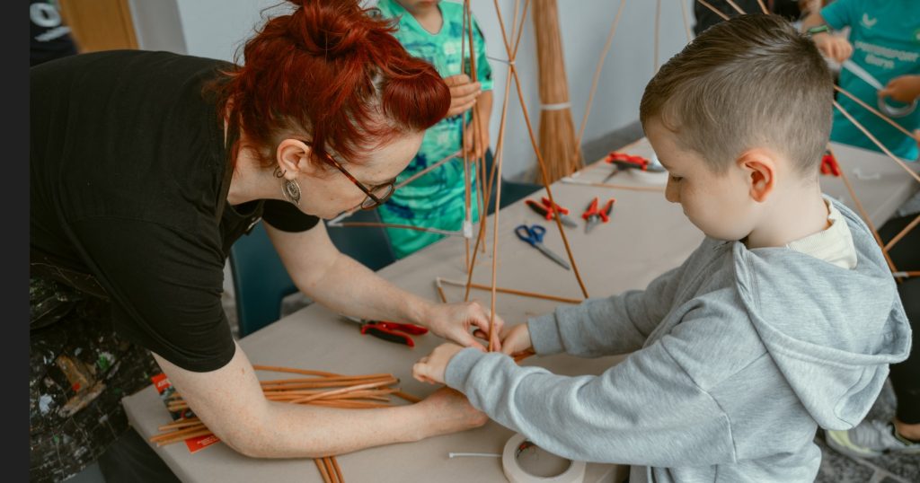 A female workshop facilitator supports a young autistic boy to construct the frame for his willow lantern.